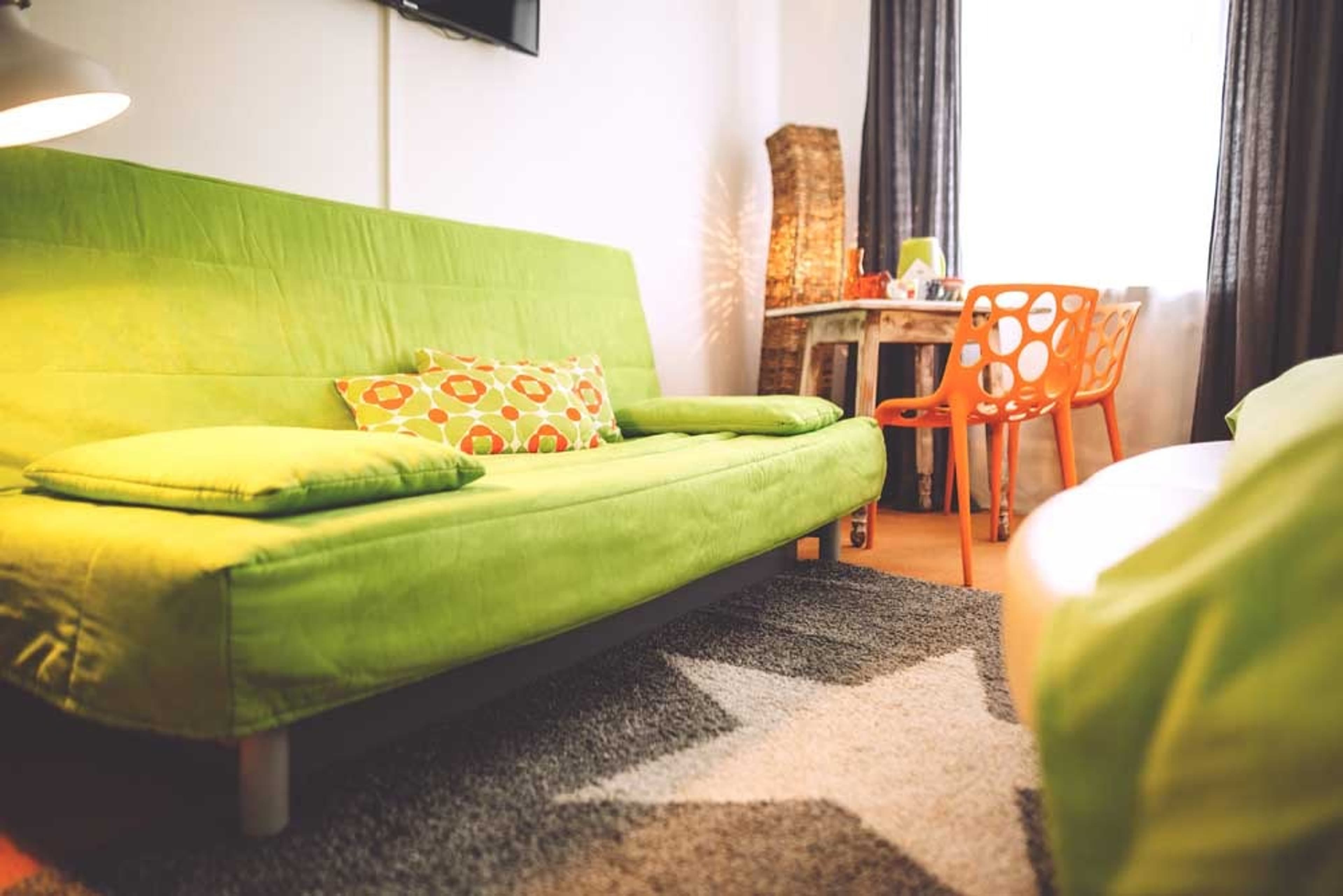 No.°1 | standard - green | no breakfast - A bright and airy bedroom featuring a double bed with white bedding and a lime green throw. A large artwork depicting sunlit trees hangs above the bed. Bedside tables with modern green lamps and a decorative ceiling lamp complete the contemporary decor.