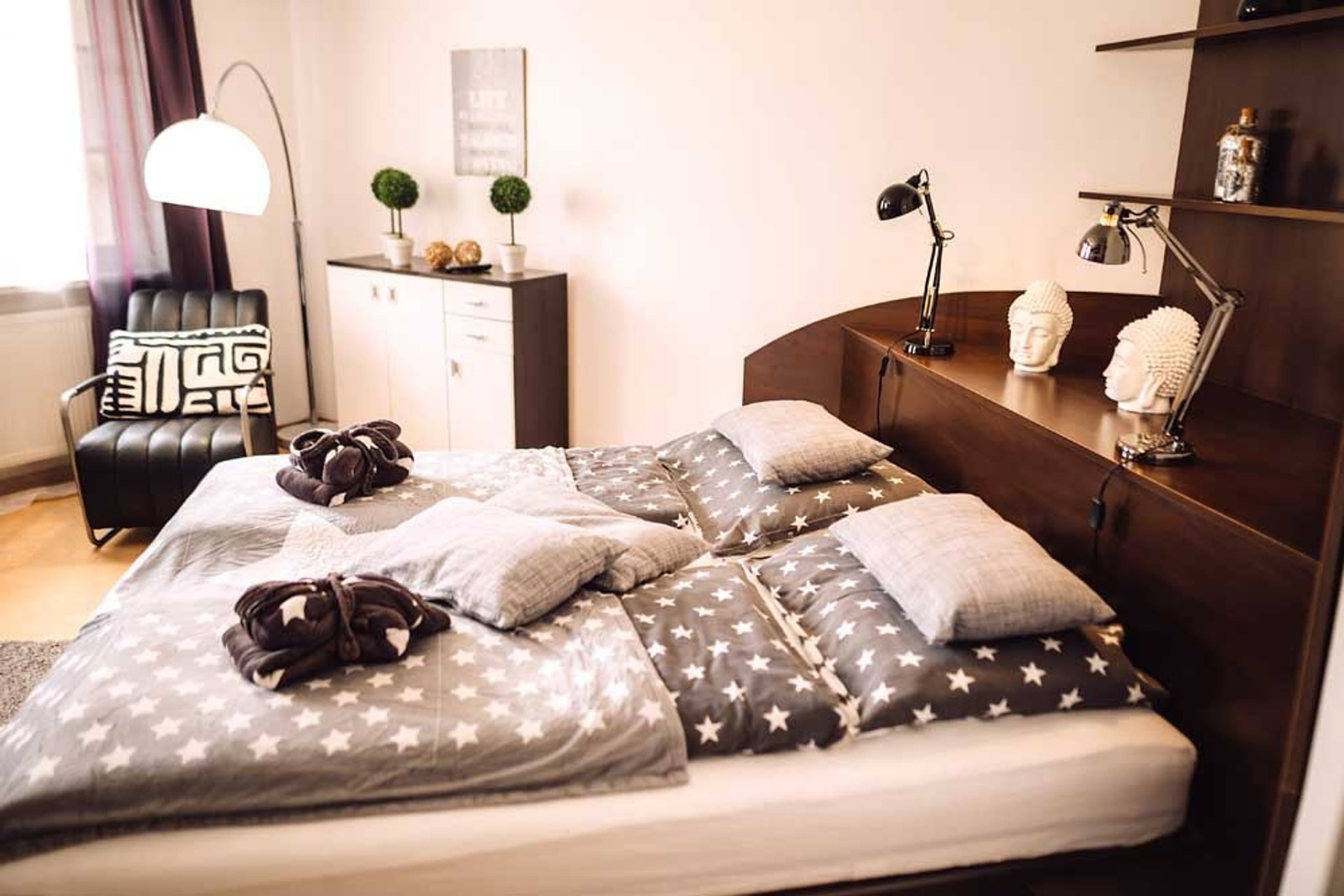 No.°3 | standard - stars | no breakfast - Bright and spacious bedroom featuring a large bed with star-patterned duvet, a grey sofa, a black armchair, and a white modern chair. Large windows with dark purple curtains let in ample natural light. A floor lamp arcs over the armchair. The room has light wooden flooring.