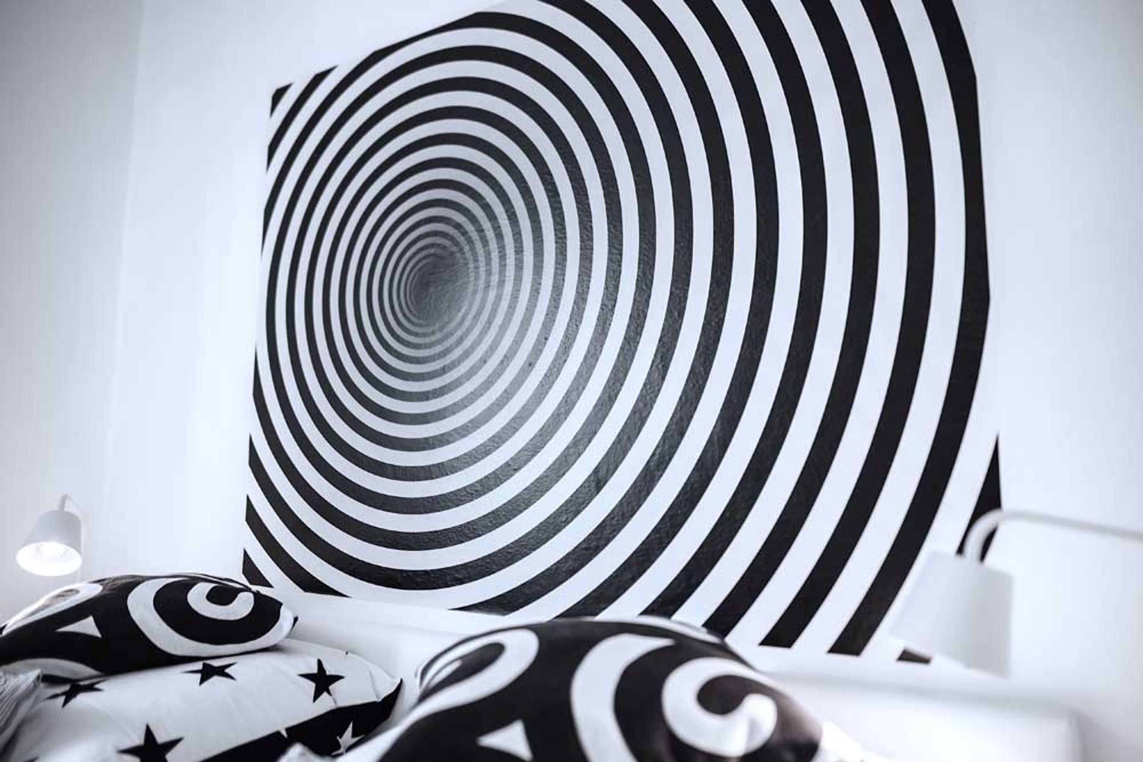 No.°4 | smart & tiny & sunny - b&w | no breakfast - Bright and modern bedroom with a large bed dressed in black and white star-patterned bedding and decorative throw pillows. A large optical illusion spiral artwork is mounted above the headboard. A window with white and black patterned curtains provides natural light, and a white chair is visible on the left.